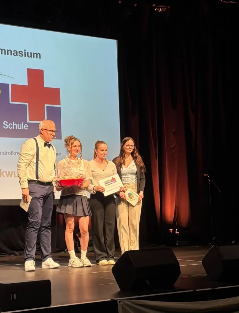 Drei Schülerinnen des GAK stehen mit ihren Urkunden auf der Bühne der Zertifizierungsfeier, daneben der Moderator.